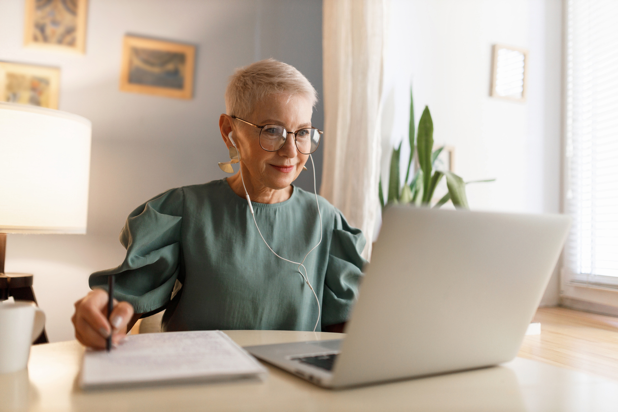 Indoor image of woman in eyeglasses watching video tutorial in wired earphones, noting down main things in her textbook, sitting near window at table desk at home